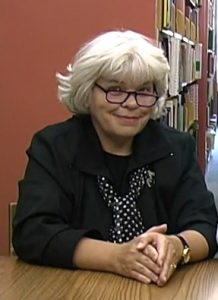 Melba Tomeo sitting at desk with library book stacks in background.